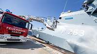 Ein Bundeswehrfeuerwehrmann steht im Korb der ausgefahrenen Leiter und sprüht Wasser auf die Bordwand eines Schiffes.