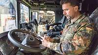 Ein militärisch gekleideter Sachverständiger der Bundeswehr sitzt hinter dem Lenkrad eines Lastwagens und notiert sich etwas. 