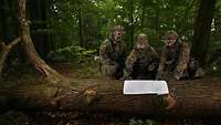 Drei Soldaten stehen vor einer Karte im Wald, die ausgebreitet auf einem umgefallenden Baumstamm liegt.