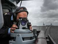 Ein Marinesoldat in blauer Arbeitsuniform blickt durch ein optisches Gerät in der Nock eines Schiffes.