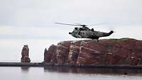Ein großer grauer Hubschrauber über dem Meer; im Hintergrund eine Insel mit roter Felsenküste.