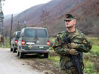 Ein Soldat steht am Straßenrand. Hinter ihm stehen zwei grüne Transporter mitde Aufschrift KFOR.