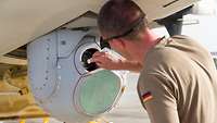 Ein Soldat im oliv-braunen T-Shirt putzt optische Linsen in einer grau-weißen Kuppel, die unter einem Flugzeugrumpf hängt