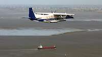 Ein zweimotoriges, blau-weißes Propellerflugzeug fliegt über Meer und Wattenmeer; direkt darunter ein Frachtschiff. 