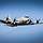 Ein hellgraues, viermotoriges Flugzeug von unten fliegt am blauen Himmel. 