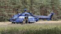 Ein blauer Hubschrauber ist an einem Waldrand gelandet, Soldaten steigen aus.