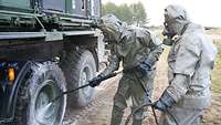 Zwei Soldaten in Schutzanzügen spritzen mit einem Hochdruckreiniger ein Fahrzeug ab