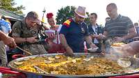 Paella schmort in einer Riesenpfanne. Ringsherum stehen Menschen mit Schüsseln und nehmen sich Reis.