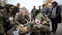 Ein verwundeter Soldat liegt angeschnallt am Boden auf einer Trage, Sanitäter kümmern sich um ihn.