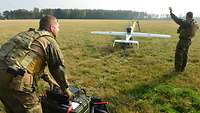 Zwei Soldaten lassen eine Drohne, die auf einer Wiese steht, starten.