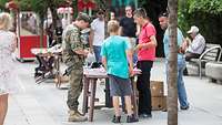Ein deutscher Soldat steht inmitten von Zivilisten an einem Straßenstand mit Verkaufswaren
