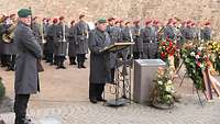 Ein General spricht, neben ihm Aufsteller mit Blumenkränzen, hinter ihm Soldaten mit roten Baretten.