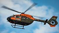 Ein grüner Hubschrauber mit orangefarbenen Aufklebern fliegt unter leicht bewölktem Himmel.