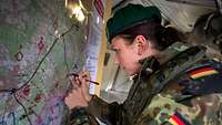 Eine junge Soldatin schreibt mit einem Stift in einem Gefechtsstand stehend an eine Lagekarte.