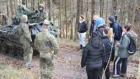 Soldaten stehen vor einem gepanzerten Fahrzeug. Eine Gruppe Jugendlicher steht im Halbkreis davor.