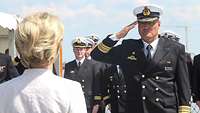 Ein Marineoffizier in dunkelblauer Uniform und weißer Tellermütze salutiert vor einer Frau in hellbeigem Blazer.