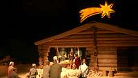 Hirten und Heilige bringen dem Christuskind ihre Gaben. Im Hintergrund der Stall mit einem Stern darüber.