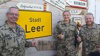 Militärpfarrer Werner Maria Heß, eine Soldatin und ein Soldat vor dem Ortsschild der Stadt Leer im Camp Castor in Mali
