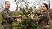 Zwei Soldatinnen schmücken einen Weihnachtsbaum. Sie schauen lächelnd in die Kamera.