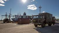 Ein Militär-Lkw steht in einem Hafen; im Hintergrund ein großes, weiß-blaues Schiff.