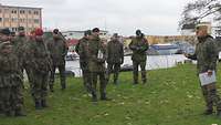 Vor Soldaten auf einer Wiese steht erklärend ein Soldat, im Hintergrund eine Stadtlandschaft.