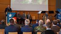 Ein Soldat steht auf einer Bühne hinter einem Rednerpult, mehrere Soldaten sitzen vor ihm im Plenum.