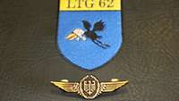 Die goldfarbene Flugzeugführerschwinge in Nahaufnahme, dahinter das Wappen des LTG 62.