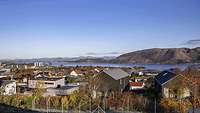 Blick über typische skandinavische Einfamilienhäuser auf einen Fjord, im Hintergrund Berge.