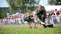 Ein Soldat hockt neben einem Hund während einer Vorführung, im Hintergrund zahlreiche Besucher hinter Absperrzäune