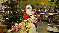 Ein Kind sitzt mit einem Glöckchen in der Hand auf dem Schoss vom Weihnachtsmann und lächelt in die Kamera.