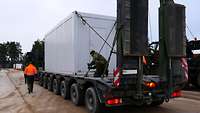Auf einem Lkw für schwere Lasten, einem sogenannten Tieflader, wird ein Container durch Soldaten fest gezurrt