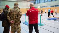 Zwei Soldaten und ein Ausbilder stehen in einer Sporthalle und schauen Soldaten beim Training zu.