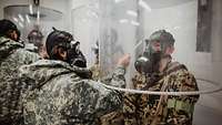 Ein Soldat steht unter einer Plastikhaube, eine amerikanische Soldatin besprüht seine ABC-Maske mit einem Prüfmittel