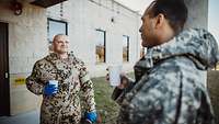 Ein deutscher und ein amerikanischer Soldat halten Trinkbecher in der Hand und unterhalten sich vor einem Gebäude