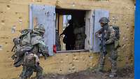 Zwei Soldaten stehen links und rechts neben einem geöffneten Fenster, im Haus sind andere Kameraden.