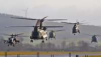 Fünf Hubschrauber verschiedener Typen fliegen bodennah über eine Landebahn.