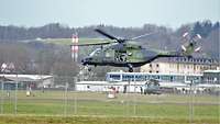 Ein Hubschrauber setzt zur Landung auf einem Flugplatz an. In der offenen Tür sitzt ein Soldat.