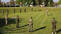 Auf einer Rasenfläche stehen Soldaten in verschiedenen Uniformen kreisförmig, mit Abstand vor einem Soldaten, der spricht.