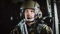 Ein Soldat mit Helm und Sprechsatz sitzt in einem Hubschrauber und schaut geradeaus.