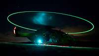 Ein Helikopter ist nachts auf einem Feld gelandet. Der mit Lichtern besetzte, drehende Rotor bildet einen Lichtkreis.