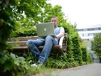 Ein junger Mann sitzt in seiner Freizeit draußen auf einer Bank vor einem Laptop.