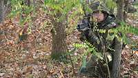 Soldat im Feldanzug kniend in einem Gebüsch, das Gewehr im Anschlag.