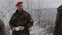 Soldat im Feldanzug vor verschneiter Landschaft, im Vordergrund Soldaten im Feldanzug von hinten.