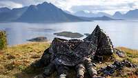 Zwei Soldaten liegen an die Umgebung angepasst auf einem grünen Hügel und blicken bei hellem Himmel auf blaues Wasser und Berge.