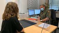 Eine junge Frau und ein Soldat sprechen an einem Bürotisch mit Plexiglasschutz
