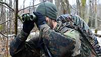 Ein Soldat mit Marschgepäck schaut in einem Wald durch ein Fernglas.