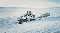 Zwei Soldaten fahren mit einem Motorschlitten mit Anhänger einen Schneehang hinunter.