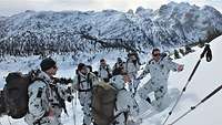 Soldaten stehen in Schneetarnanzügen knietief im Schnee an einem Hang, hinter ein Gebirgsmassiv.
