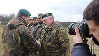 Ein Soldat gratuliert einem älteren Soldaten mit Vollbart und Brille. Ein Journalist fotografiert.