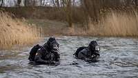 Zwei Soldaten in Taucherausrüstung stehen nahe einem mit Schilf bewachsenen Ufer im Wasser.